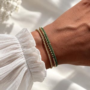 Könnte beinhalten: Zwei zarte Armbänder am Handgelenk. Ein Armband besteht aus kleinen, polierten goldfarbenen Kugeln. Das zweite Armband besteht aus winzigen, facettierten grünen Edelsteinperlen, die mit kleinen goldfarbenen Zwischenperlen kombiniert sind.