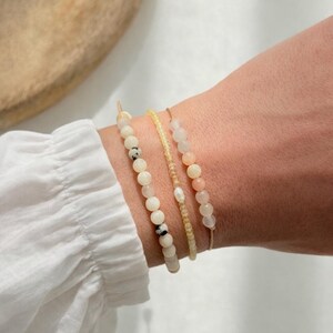 Könnte beinhalten: Drei Armbänder mit Perlen in Creme-, Gelb- und Pfirsichtönen werden an einem Handgelenk getragen. Die Armbänder weisen unterschiedliche Perlengrößen und -strukturen auf, darunter gesprenkelte und glatte Perlen. Das Handgelenk befindet sich neben einem weißen Ärmel.