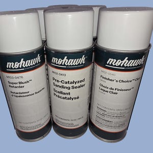 May include: Three cylindrical aerosol spray cans with white caps and white labels. The labels feature the Mohawk brand name and product names: Super Blush Retarder, Pre-Catalyzed Sanding Sealer, and Finisher's Choice Clear. Each can contains 13 oz.