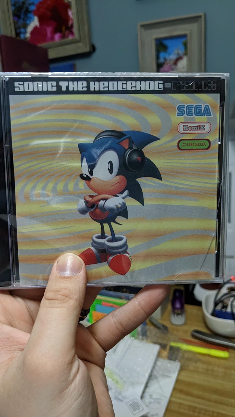 May include: A Sonic the Hedgehog Remix CD case featuring the character Sonic with headphones. The case has a yellow and orange background with the Sega logo and the text "Sonic the Hedgehog - Remix".