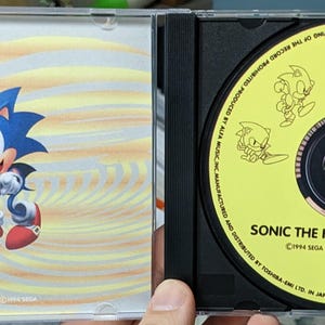 May include: A Sonic the Hedgehog CD case. The case features a blue Sonic character on a yellow and orange background. The CD itself is yellow with Sonic illustrations and the text "SONIC THE HEDGEHOG-REMIX".
