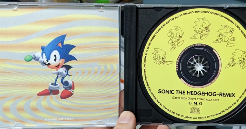 May include: A Sonic the Hedgehog CD case. The case features a blue Sonic character on a yellow and orange background. The CD itself is yellow with Sonic illustrations and the text "SONIC THE HEDGEHOG-REMIX".