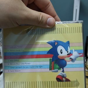 May include: A CD case featuring the character Sonic the Hedgehog. The case has a colorful striped background with song titles listed. The Sonic character is blue with white gloves and red shoes. The text "Love you Sonic" is visible.