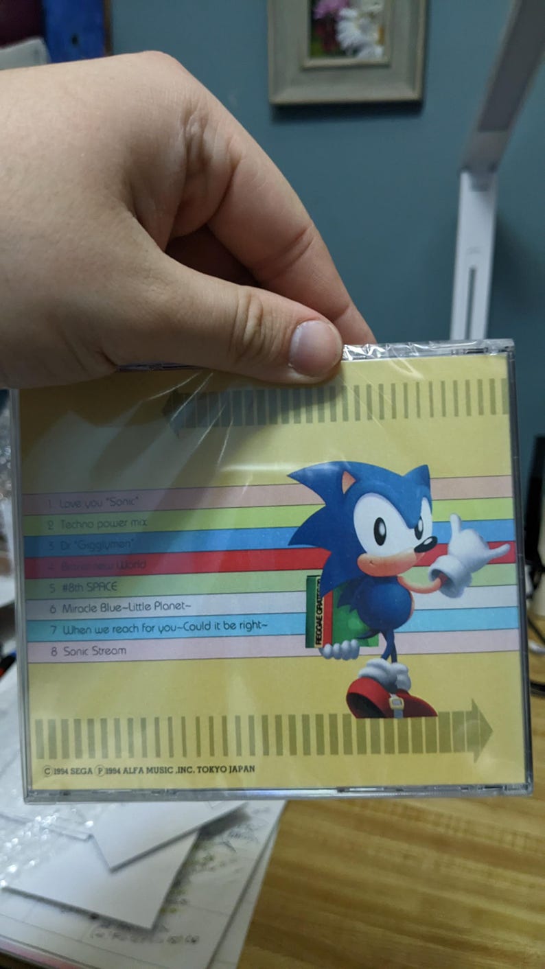 May include: A CD case featuring the character Sonic the Hedgehog. The case has a colorful striped background with song titles listed. The Sonic character is blue with white gloves and red shoes. The text "Love you Sonic" is visible.