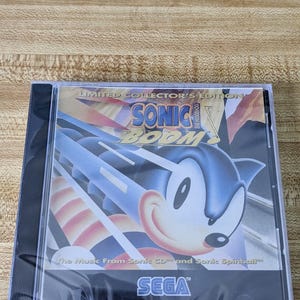 May include: A Limited Collector's Edition CD case for Sonic Boom. The cover features Sonic the Hedgehog in a dynamic pose with the text "SONIC BOOM" and "The Music From Sonic CD and Sonic Spinball" and the SEGA logo.