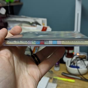 May include: A CD case for "Sonic the Hedgehog - Remix" is held in a hand. The spine of the case displays the title and a colorful striped design. The text "ALCA-5023 ALFA MUSIC, INC." is also visible.