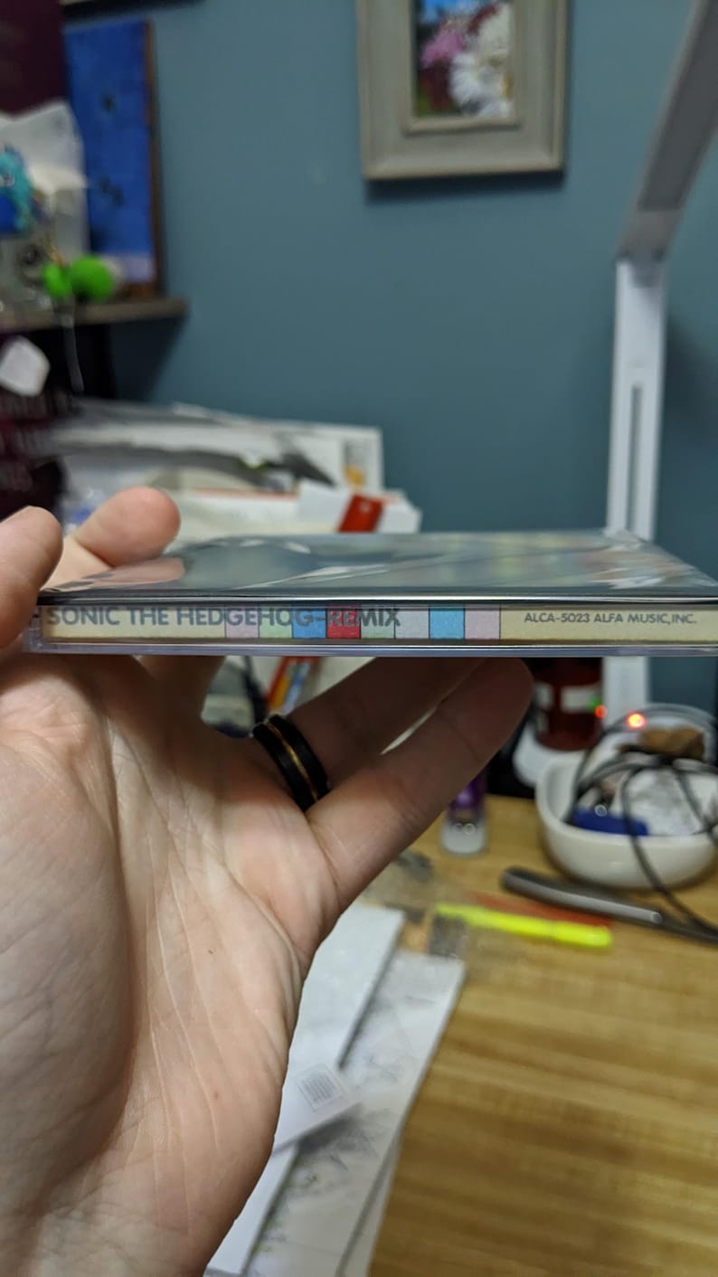 May include: A CD case for "Sonic the Hedgehog - Remix" is held in a hand. The spine of the case displays the title and a colorful striped design. The text "ALCA-5023 ALFA MUSIC, INC." is also visible.