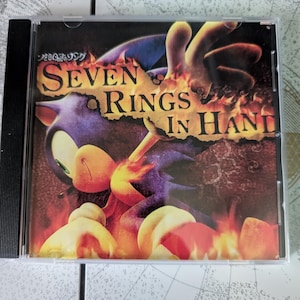 May include: A CD case featuring the Sonic the Hedgehog character with the text "SEVEN RINGS IN HAND". The artwork depicts Sonic in a fiery setting with flames and a dark background. The CD case is in a clear plastic case.