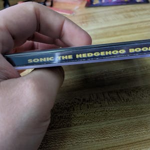 May include: A clear plastic case with the text "SONIC THE HEDGEHOG BOOM" in gold lettering on a black background. The case is held in a hand and is resting on a wooden surface.