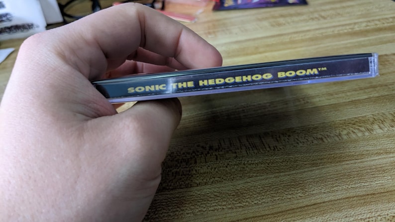 May include: A clear plastic case with the text "SONIC THE HEDGEHOG BOOM" in gold lettering on a black background. The case is held in a hand and is resting on a wooden surface.