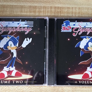May include: Two Sonic the Hedgehog 30th Anniversary Symphony CD cases. The covers feature Sonic in a tuxedo conducting with a baton. The text reads 30th Anniversary Symphony, with Volume One on the right and Volume Two on the left, set against a black background with musical notes and glowing light effects.