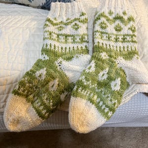 May include: A pair of hand-knitted socks with a white and green pattern. The socks feature a white toe and cuff, with a green and white geometric design on the foot and leg. The socks are made of a soft, textured yarn.