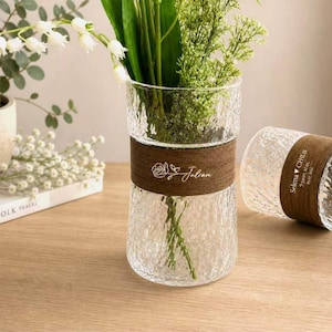 May include: Two clear glass vases with a textured, hammered surface. Each vase features a dark brown wooden band around the center. One vase is upright holding green stems, while the other lies horizontally, displaying the text Selena ♥ CHLAI.