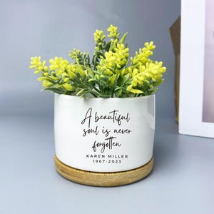 以下が含まれることがあります： 木製の台座が付いた白いセラミック製の植木鉢。黄色と緑色の造花が入っています。「A beautiful soul is never forgotten」というテキストと「KAREN MILLER 1967-2023」という名前が書かれています。