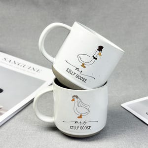 May include: Two white ceramic mugs stacked, each with a whimsical goose illustration. One goose wears a top hat and bow tie, the other a veil. The mugs read "Mr. Silly Goose" and "Mrs. Silly Goose".