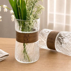 May include: Two clear glass vases with a textured, crinkled surface and a wide, dark brown wooden band around the center. The band features the engraved letters B and K, a small floral icon, and the date 08.11.2025. One vase holds green stems.