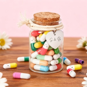 May include: A glass jar filled with colorful capsule-shaped candies, some with smiling faces, and a cork lid. A heart-shaped charm with "S & B" hangs from the jar. Daisies and scattered candies are on a wooden surface.