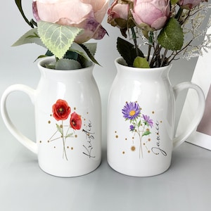 May include: Two white ceramic vases with handles, each adorned with floral designs and names. One vase features red poppies and the name "Kristin", while the other showcases purple flowers and the name "Janice". Both vases hold flowers.
