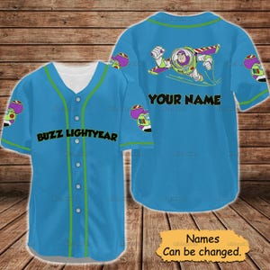 May include: A light blue baseball jersey featuring Buzz Lightyear graphics. The front displays "BUZZ LIGHTYEAR" in black, while the back has a Buzz Lightyear image and the text "YOUR NAME". Green accents trim the collar, sleeves, and button line. The jersey is a fun, themed apparel item.