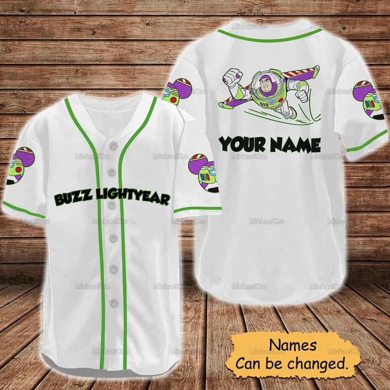 May include: White baseball jersey with green trim and the words "BUZZ LIGHTYEAR" across the front. The back features a Buzz Lightyear graphic and the text "YOUR NAME". The jersey has Buzz Lightyear graphics on the sleeves. The text "Names Can be changed" is on a yellow sign.
