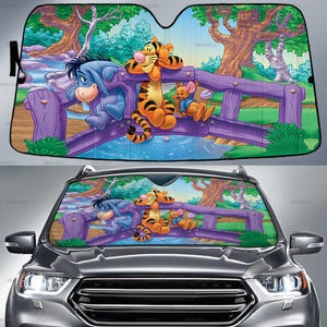 Puede incluir: Parasol para coche con una colorida ilustración de los personajes de Winnie the Pooh. Ígor, Tigger y Rito se representan en un puente morado sobre un arroyo azul, con un fondo de bosque. El parasol está diseñado para el parabrisas de un coche.