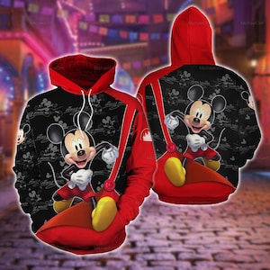 Puede incluir: Una sudadera con capucha negra y roja que presenta un patrón repetitivo de Mickey Mouse sobre un fondo negro. La parte delantera y trasera muestran un gráfico grande y colorido de Mickey Mouse con pantalones cortos rojos y zapatos amarillos.