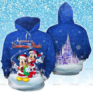 Könnte beinhalten: Ein blauer Kapuzenpullover mit einer Grafik von Mickey und Minnie Mouse in Weihnachtsmannkostümen, die einen grünen Strumpf halten. Der Text lautet Believe In CHRISTMAS MAGIC. Die Rückseite zeigt ein großes Schloss in einer Schneelandschaft.