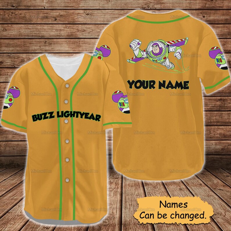 May include: Mustard yellow baseball jersey featuring Buzz Lightyear graphics. The jersey has green trim and the text "BUZZ LIGHTYEAR" on the front. The back has a Buzz Lightyear graphic and the text "YOUR NAME". The text "Names Can be changed" is also visible.