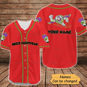 May include: Red baseball jersey featuring Buzz Lightyear graphics. The front displays "BUZZ LIGHTYEAR" in black, with green trim and buttons. The back has a Buzz Lightyear graphic and space for a name. The sleeves have character details.