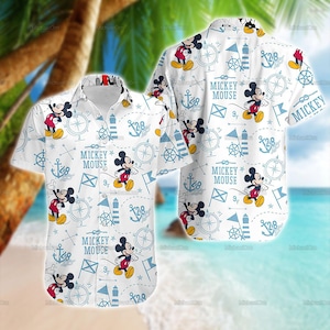May include: White button-up shirt featuring a repeating pattern of Mickey Mouse, anchors, compasses, and nautical flags in blue. The words "Mickey Mouse" are printed on the shirt. The shirt has short sleeves.