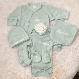 Peut inclure: Un ensemble de vêtements pour bébé vert sauge comprenant une grenouillère à manches longues, un body à manches courtes, un bonnet, un bavoir et une paire de moufles. Chaque pièce est brodée du prénom Imran en blanc, sauf les moufles avec un J.