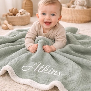 Op de afbeelding: Een saliegroene babydeken met witte sherpa-afwerking en de naam "Atkins" geborduurd in witte letters. De deken is gedrapeerd over een zacht, neutraal gekleurd tapijt. Een lachende baby staat op de foto.
