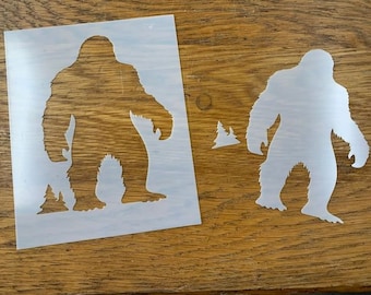Peut inclure: Deux pochoirs en vinyle blanc de Bigfoot, l'un plus grand que l'autre, avec un petit pochoir d'arbre en arrière-plan. Les pochoirs sont sur une surface en bois.