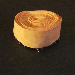 May include: A rolled-up, cylindrical bundle of orange fabric. The fabric is tightly wound, creating a spiral pattern on the top surface. The edges of the fabric have loose, frayed threads. The object is set against a dark background.