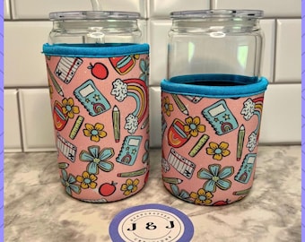 May include: Two clear glass tumblers with pink and blue school-themed neoprene can coolers. The coolers feature a pattern of pencils, rulers, notebooks, and flowers. The text "J & J HANDCRAFTED CREATIONS" is at the top of the image.