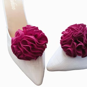 May include: White pointed-toe mules with a textured pattern, each adorned with a ruffled, deep magenta fabric flower. The shoes have a minimalist design, perfect for adding a touch of elegance to any outfit.