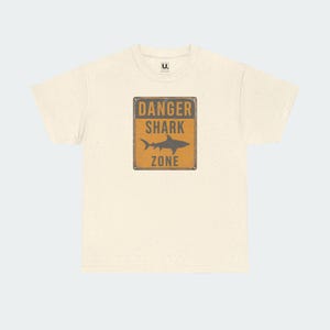 A white t-shirt with a graphic design featuring a warning sign that says "DANGER SHARK ZONE".