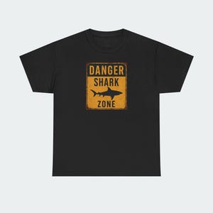 A black t-shirt with a graphic design featuring a shark silhouette and the text "DANGER SHARK ZONE" printed on it.