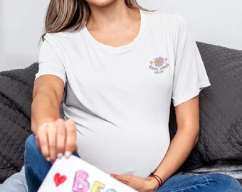 Op de afbeelding: Een wit T-shirt met korte mouwen en een kleine afbeelding op de linkerborst met een lachende bloem en de tekst "COOL MOM CLUB" in pastelkleuren. Het T-shirt wordt gecombineerd met een blauwe spijkerbroek en vastgehouden met een "BEST MOM"-bord.