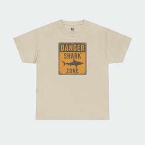 A beige t-shirt with a graphic design featuring a shark and the text "DANGER SHARK ZONE" printed on it.