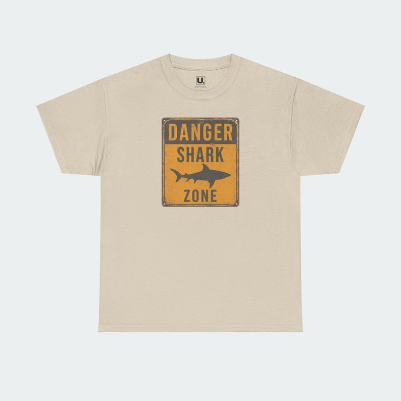A beige t-shirt with a graphic design featuring a shark and the text "DANGER SHARK ZONE" printed on it.