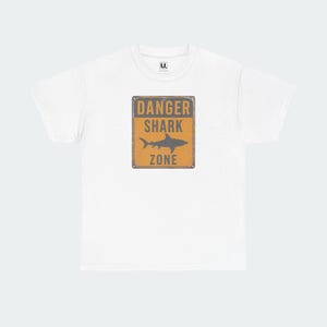 A white t-shirt with a graphic design featuring a shark and the text "DANGER SHARK ZONE" printed on it.
