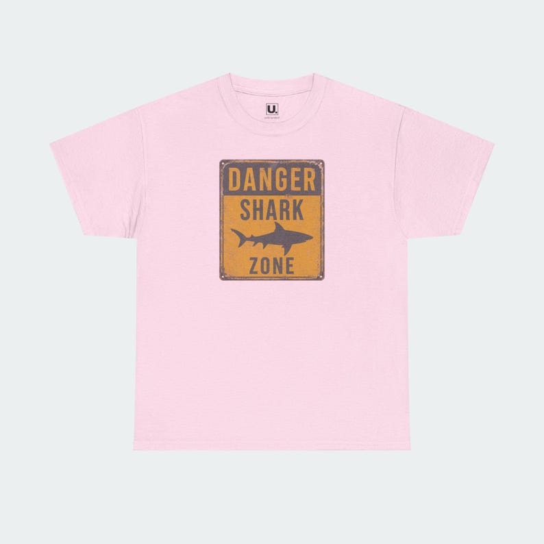 A pink t-shirt with a graphic design featuring a shark and the text "DANGER SHARK ZONE" printed on it.