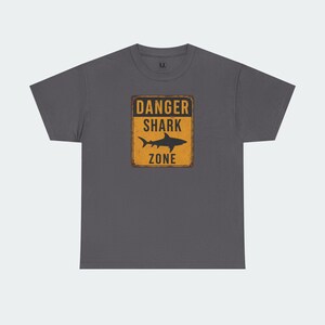 A gray t-shirt with a graphic design featuring a shark silhouette and the text "DANGER SHARK ZONE" printed on it.