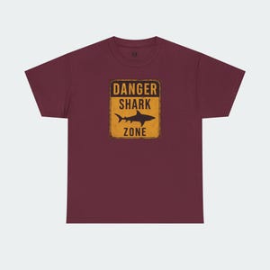 A burgundy-colored t-shirt with a graphic design depicting a shark and the text "DANGER SHARK ZONE" printed on it.