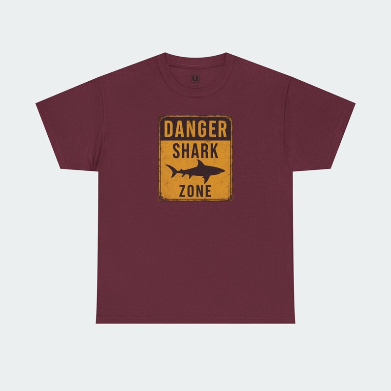 A burgundy-colored t-shirt with a graphic design depicting a shark and the text "DANGER SHARK ZONE" printed on it.