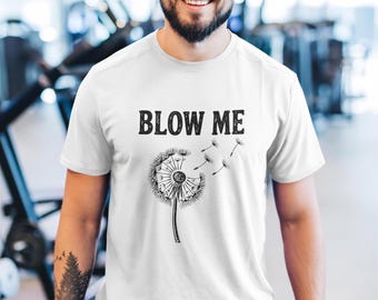 Op de afbeelding: Een wit T-shirt met korte mouwen en een zwarte afbeelding van een paardenbloem waarvan de zaadjes in de wind wegwaaien. De tekst "BLOW ME" is in een vet, zwart, verweerd lettertype boven de paardenbloemillustratie op de borst gedrukt.