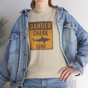 A person wearing a denim jacket and a t-shirt with a "Danger Shark Zone" graphic, posing in front of a white wall.