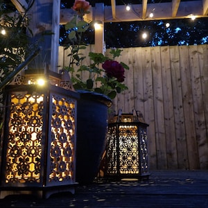 May include: Two decorative lanterns with intricate cut-out patterns are illuminated, casting a warm glow. A dark blue flower pot with a rose bush is between the lanterns. The scene is set outdoors with a wooden fence and string lights.