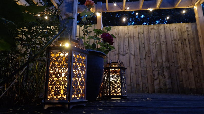 May include: Two decorative lanterns with intricate cut-out patterns are illuminated, casting a warm glow. A dark blue flower pot with a rose bush is between the lanterns. The scene is set outdoors with a wooden fence and string lights.